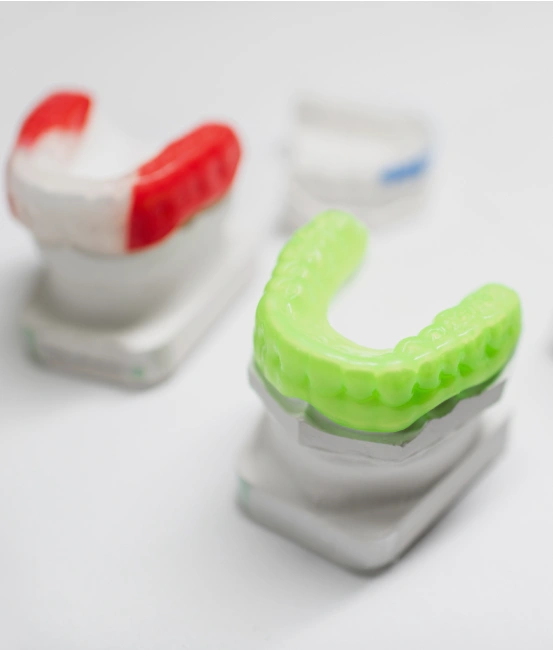 A bright green custom dental mouthguard sitting on a stone mold model of teeth, with a red-and-white guard visible in the background.